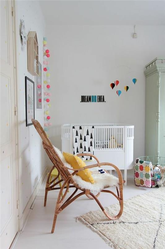 kūdikio mergaitės kambario apdaila, austa rotango kėdė šviesios spalvos, kūdikio kambario stalas, ovalo formos kūdikio lova, pastelinės spalvos kilimėliai