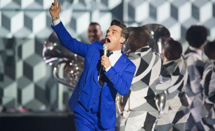 koyu mavi takım elbise ceketi, konserde Robbie Williams, beyaz gömlek, siyah kravat, koyu mavi yelek, büyük sahne