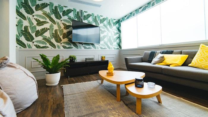 doğru televizyon nasıl seçilir, egzotik duvar kağıdı arka planında yüksek bir duvarda ayarlanabilir TV sehpası ile oturma odası dekorasyonu örneği, gri kanepe, sarı minderler, ahşap açılır masalar