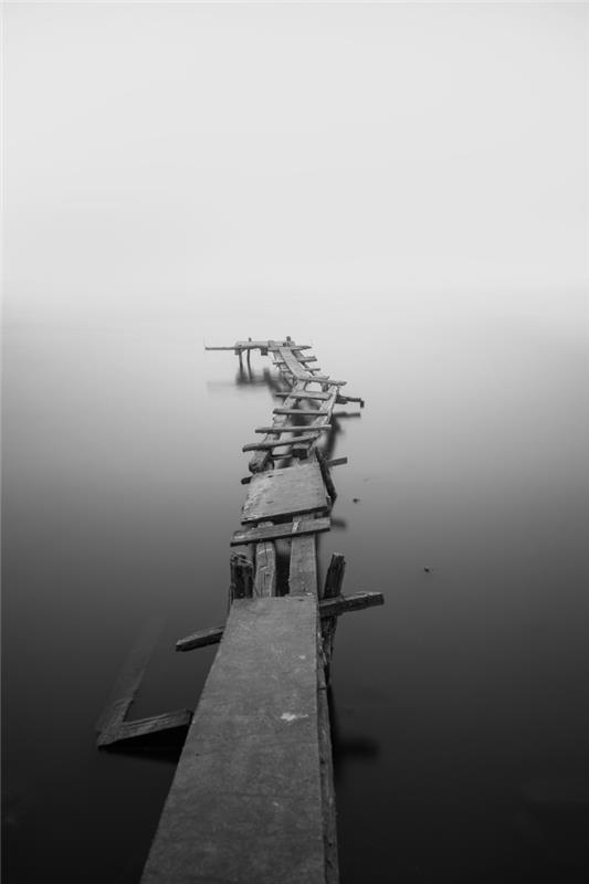 črno -bela podoba starega lesenega pomola, obdanega z mirno morsko vodo