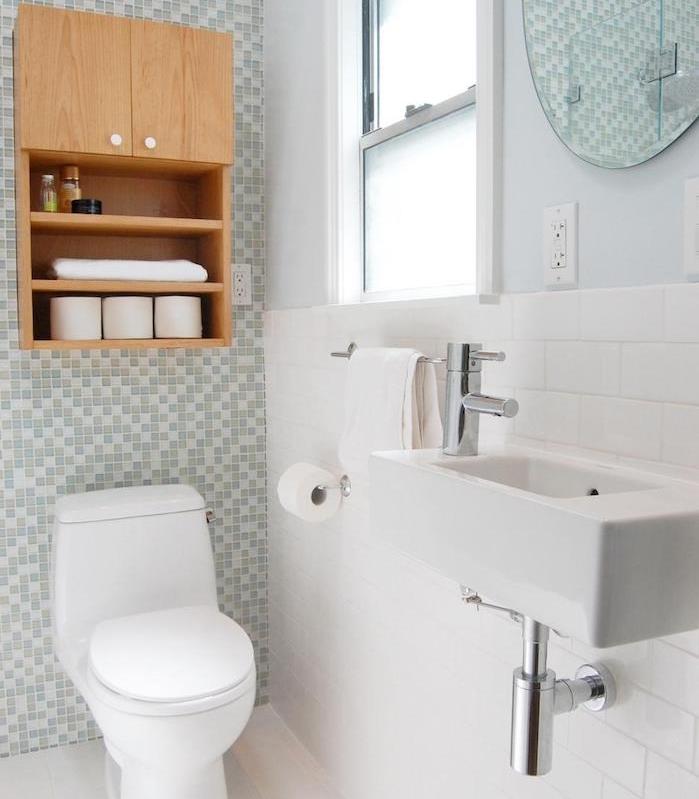 gri ve beyaz kiremitli bir duvar ve beyaz fayansların geri kalanı, beyaz lavabo ve tuvalet, yuvarlak ayna, tuvaletin üstünde dolap bulunan banyo düzeni