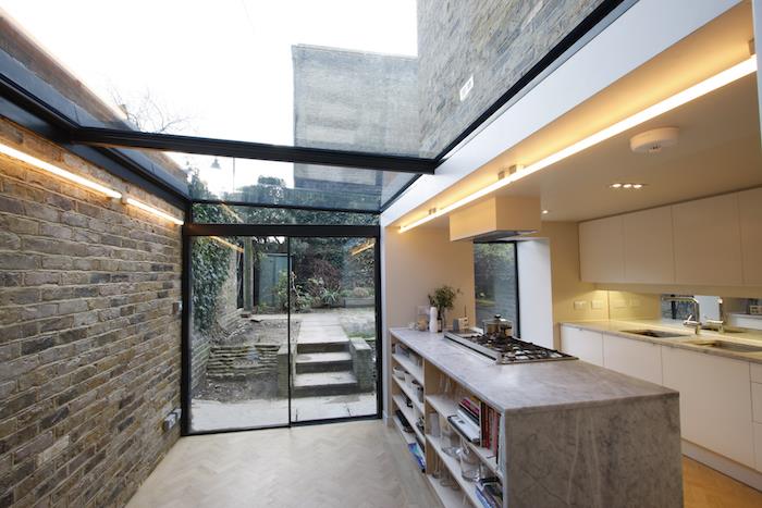 namų išplėtimo išdėstymas baltoje virtuvėje su pilko marmuro centrine sala, šviesiu parketu, išoriniu stikliniu stogu ir LED apšvietimu