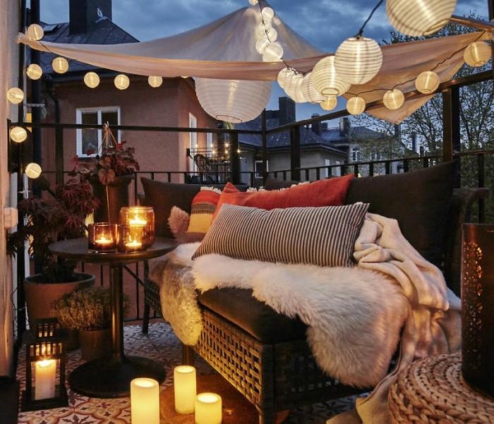 majhna romantična postavitev balkona s kavčem, okrašenim s kariranimi in okrasnimi blazinami, več svečniki in svečami, senčnikom in kroglico