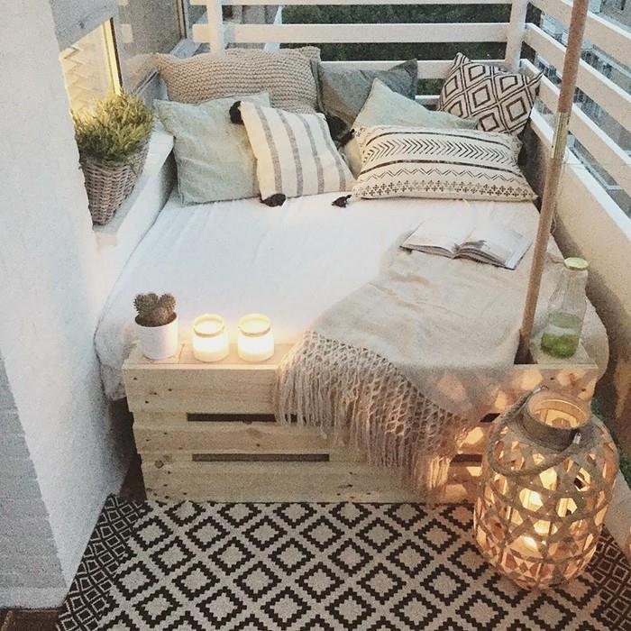 maža padėklų medinė sofa su pilka ir smėlio spalvos čiužiniu bei dekoratyvinėmis pagalvėlėmis, pilka ir juoda kiliminė danga, balkono žvakidės ir žvakės