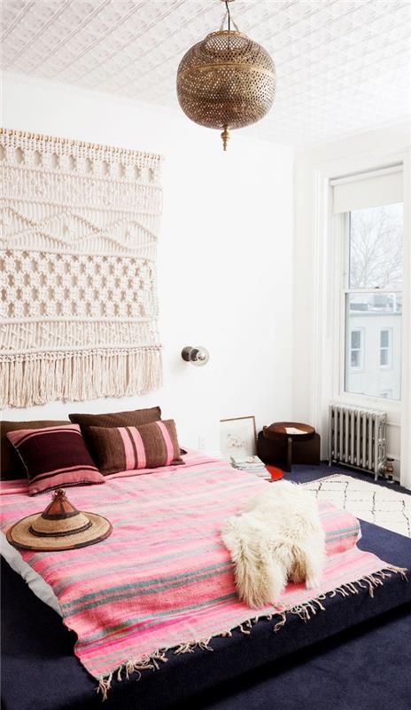 prašmatnus bohemiškas dekoras su pakabinamu makramežiu ir šviesiai pilkos ir rausvos spalvos dryžuotu pledu su pakraščiais, lovos modelis ant grindų, padengtas languotomis ir dekoratyvinėmis pagalvėlėmis