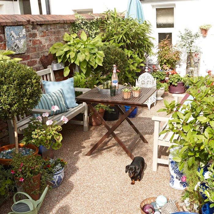 mažas sodo išdėstymas, aptio sodo baldai, baltas suoliukas ir sulankstomas medinis stalas, mėlynos dekoratyvinės pagalvėlės, kelios gėlės ir krūmai, pasodinti gėlių vazonuose