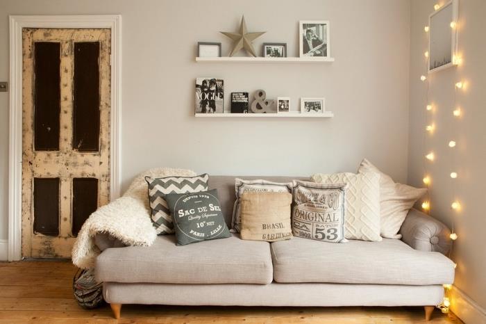 maža poilsio erdvė su jaukia smėlio spalvos sofa, padengta to paties tono toninėmis dekoratyvinėmis pagalvėlėmis, ir ant sienos kabančia lengva girliandos puošmena