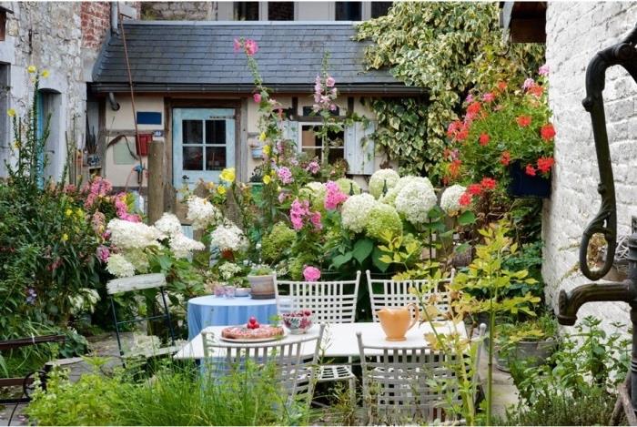 mažas gėlių sodas su daugybe gėlių ir žydinčių krūmų bei vijoklinių augalų, lauko valgomojo zona su baltu stalu ir kėdėmis