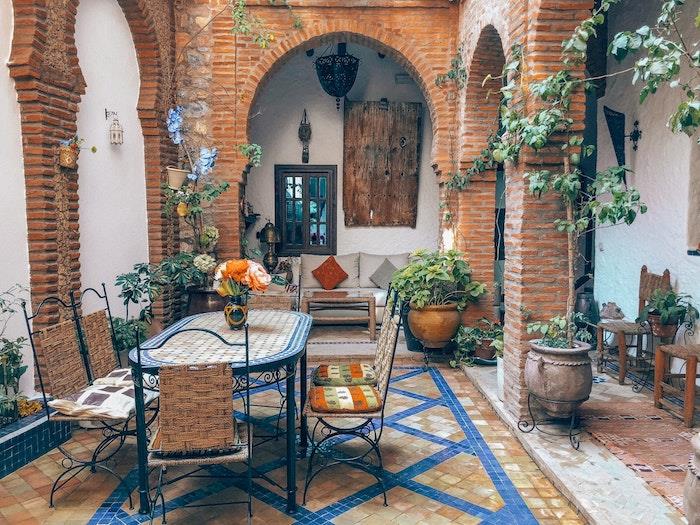 Yerde mozaik üzerine masa ve sandalyelerden oluşan yemek alanı, gri masa ve kanepe, bohem şık tarzı bahçe dekorundan oluşan ferforje bahçe mobilyası örneği