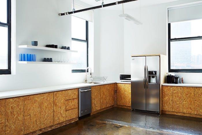 įrengta virtuvė su žemais mediniais baldais ir baltu stalviršiu, baltos lentynos, nerūdijančio plieno prietaisai, cinko grindys