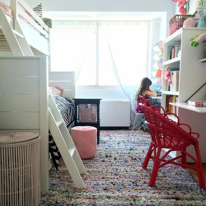 večbarvna preproga, bela lesena podstrešna postelja z lestvijo, tkani rdeči stoli, bela knjižna omara, bež košara, roza stolček