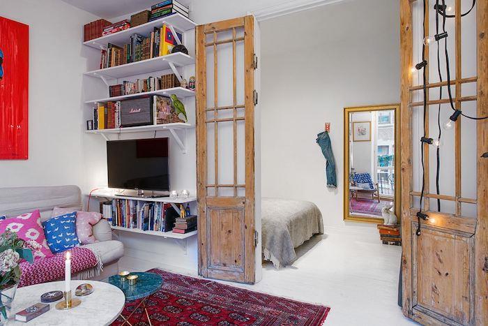 miegamasis su svetaine su pilka sofa, papuošta spalvingomis pagalvėmis, raudonu rytietišku kilimu, marmuriniu kavos staliuku, medinėmis durimis su senomis langinėmis, elegantiška bohemiška studija