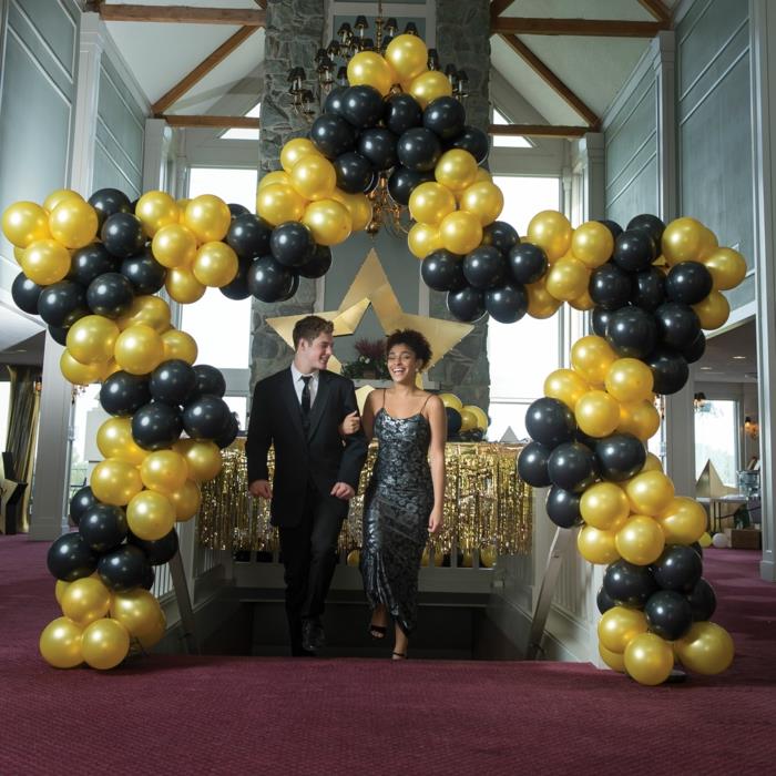 žvaigždžių balionų dekoravimas juodais ir geltonais balionais, jauna pora, vyras su kostiumu ir moteris su oficialia suknele, didelė lubų lempa