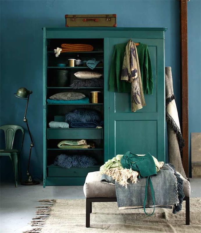 benzinu mėlynai dažyta spintelė, atsirėmusi į tamsiai mėlyną sieną su smaragdo žalios spalvos dekoratyviniais akcentais, žalia kėdė ir lempa, senovinė bagažinė