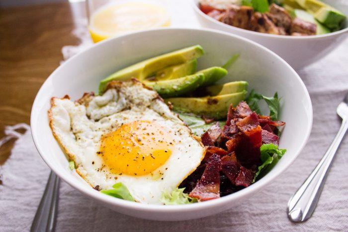 daug baltymų turintis pusryčių dubuo, žalios salotos, keptas kiaušinis, šoninė, šoninė ir avokado griežinėliai