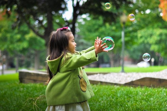 Maža mergaitė, žaidžianti su muilo balionais, mažos mergaitės šukuosena, pynimo šukuosena, lengva šukuosenos pamoka, žalias kailis, žalias sodo pavasaris