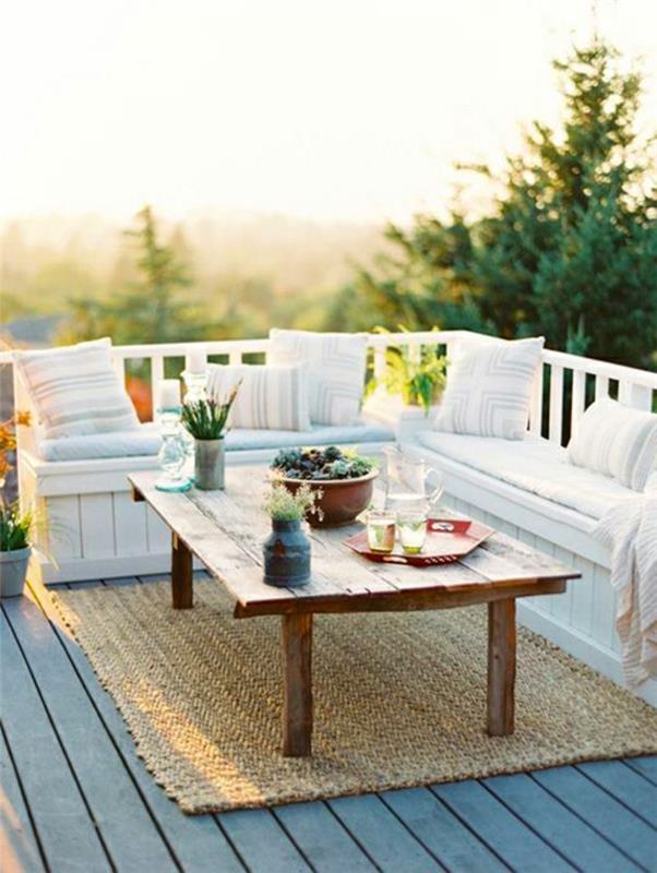 lauko kampinė balta medžio sofa, dekoruota baltomis pagalvėlėmis, pilka kompozicinės medienos terasa, smėlio spalvos kilimas, kaimiškas kavos staliukas