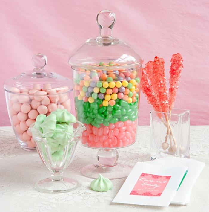 bir şeker çubuğunun nasıl organize edileceğine dair fikir, pastel renkler, renkli şekerli bademler, lolipoplar, yeşil bezeler, pembe arka plan, beyaz masa örtüsü, cam şeker kutusu