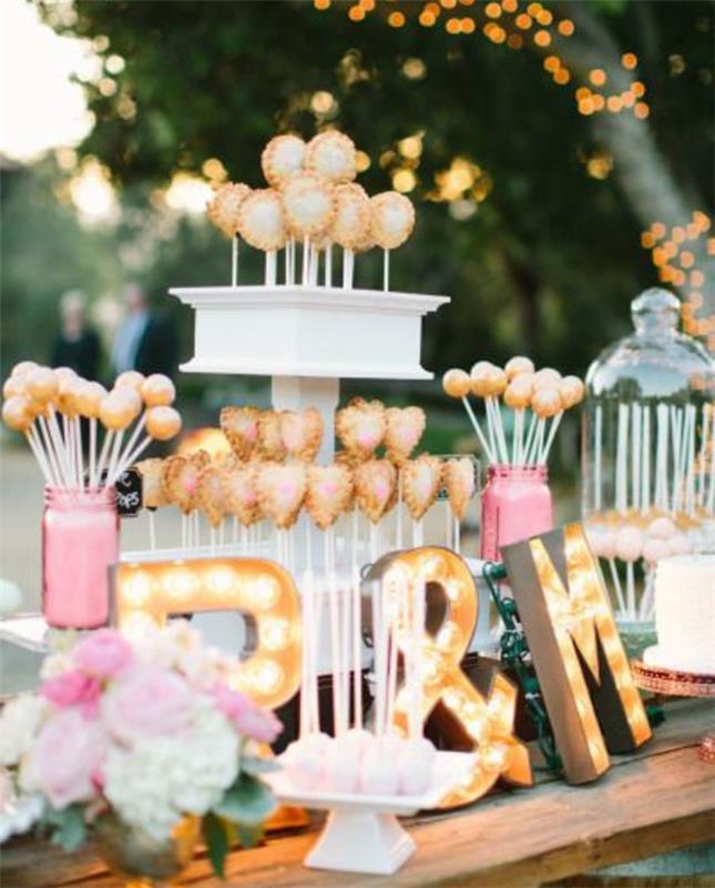 sladkarije v roza, beli in zlati barvi, lizike za torte, šoto rustikalna miza, cvetlični šopek, svetle črke, poroka na prostem