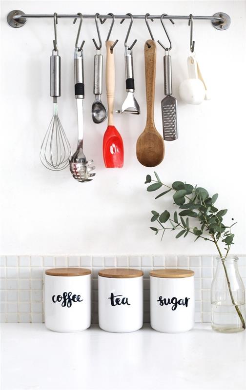 metalinis sieninis baras su kabliukais virtuvės reikmenims pakabinti, trys balti stiklainiai su kamštiniais dangteliais kavai, arbatai ir cukrui