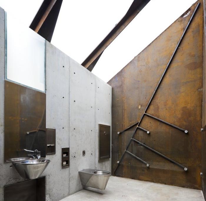 ideje za dekoracijo kopalnice, stranišče v industrijskem slogu, z betonskimi in zarjavelimi kovinskimi stenami, navadnim srebrnim umivalnikom in WC školjko, nagnjenim stropom s kovinskim nosilcem, ki podpira čisto stekleno površino