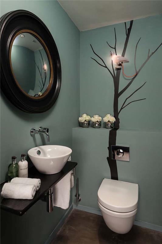 ideje za dekoracijo kopalnice, stranišče s stenami v modri barvi račjega jajca, eno steno krasi stenska poslikava iz črnega drevesa brez listov, okroglo ogledalo z velikim temnim lesenim okvirjem visi na drugi vidni steni