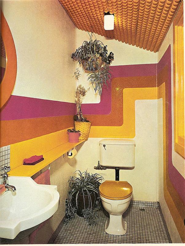 stranišče iz retro šestdesetih let prejšnjega stoletja, siva mozaična tla, retro umivalnik, straniščni sedež z lesenim pokrovom, stene z rožnatimi, oranžnimi in rumenimi upogibnimi črtami, več lončnic, oranžno teksturiran strop