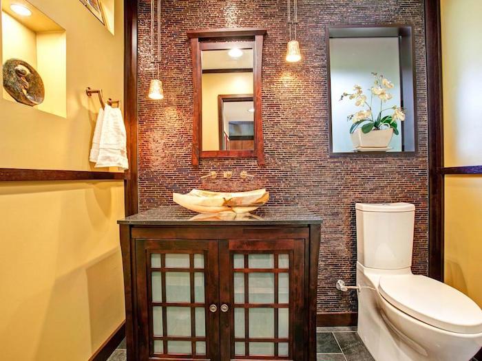 ideje za dekoracijo kopalnice, stranišče z dvema rumenima stenama in tretjo steno, okrašeno z rjavim mozaikom, lesenimi tramovi in ​​obrobami, velika lesena omara z nenavadnim umivalnikom, sodobno belo stranišče