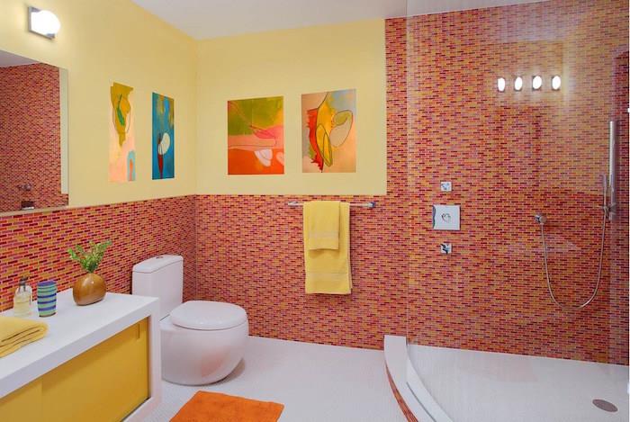 ideje za kopalnico, velika soba z veliko stekleno tuš kabino, belo -rumeno omarico, okroglo moderno belo straniščno školjko, stene napol pokrite z rdečim, oranžnim in rumenim mozaikom, druga polovica pobarvana v rumeno, veliko ogledalo in slike na stenah