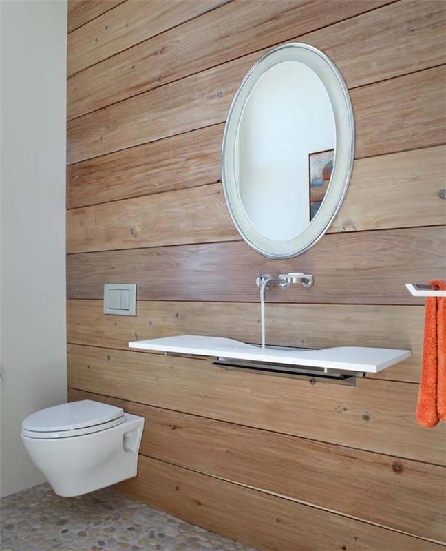 prenova kopalnice, prostorno stranišče z eno steno, prekrito z lesenimi deskami, druga vidna stena je bela, okroglo ogledalo s srebrnim okvirjem, beli minimalistični moderni umivalnik, prodnata tla in navadna bela WC školjka