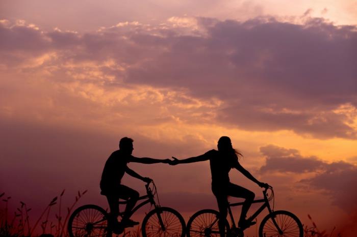 Bisikletlerinde aynı anda el ele tutuşan kadın ve erkeğin çift siluetinin havalı fotoğrafı, pembe-mor gökyüzü