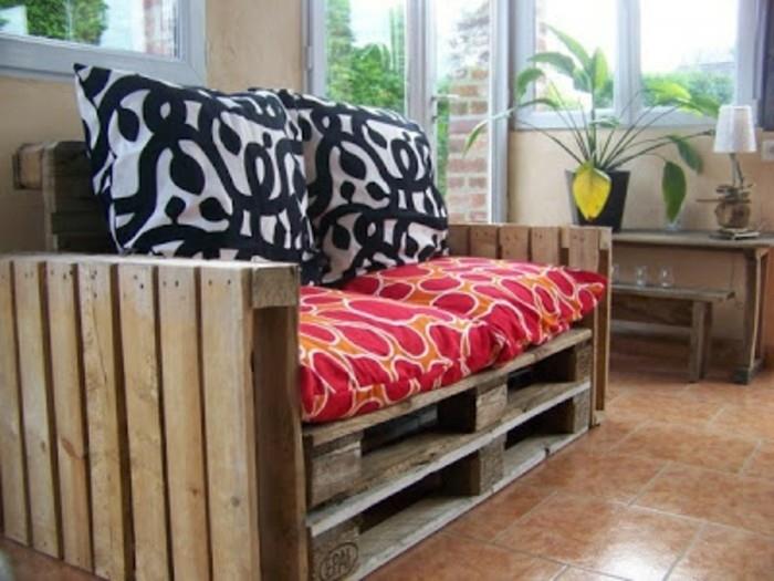 pagalvėlės juodos ir baltos, oranžinės ir raudonos spalvos, dekoruojančios „pasidaryk pats“ sofą, pagamintą iš medinių padėklų, šviesiame kambaryje su nedideliu stalu ir vazoniniu augalu