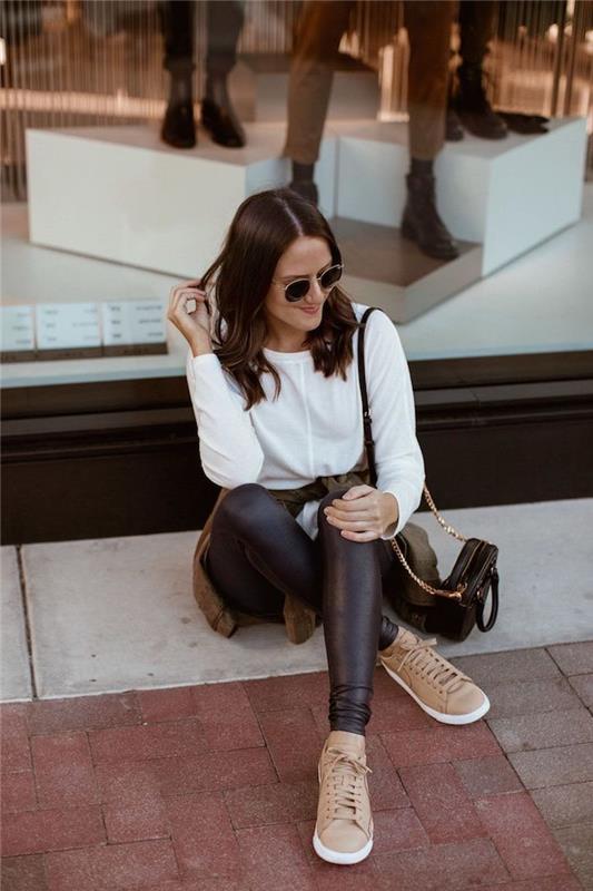 ženska, ki sedi na ulici, trenutni modni trendi, nosi črne usnjene nogavice in belo bluzo, bež superge in črno torbo