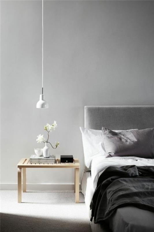 minimalistinis miegamasis su šviesiai pilkomis sienomis, nedidelis medinis naktinis stalas, pilka lova su baltos, šviesiai ir tamsiai pilkos spalvos lovatiesėmis