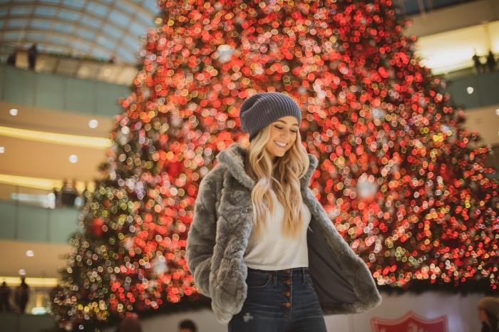 linksmų Kalėdų nuotrauka su mergina, apsirengusia džinsais ir dirbtinio kailio kailiu prekybos centre, papuoštame milžiniška egle