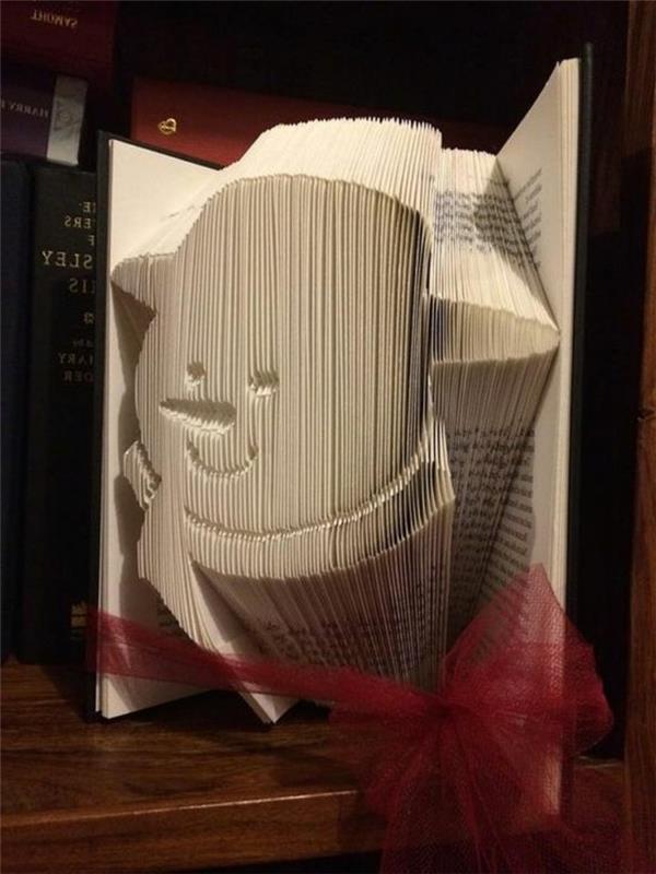 vzorci zlaganja knjig, odprta knjiga s črnimi platnicami, vezana s prozornim rdečim trakom, ki prikazuje nasmejano glavo snežaka, narejeno iz prepognjenih strani