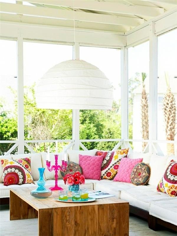 kapalı veranda fikirleri, parlak renkli dekoratif objelerin olduğu ahşap masa, farklı şekil ve renklerde birçok minder ile büyük beyaz ve kahverengi köşe kanepe, büyük beyaz avize