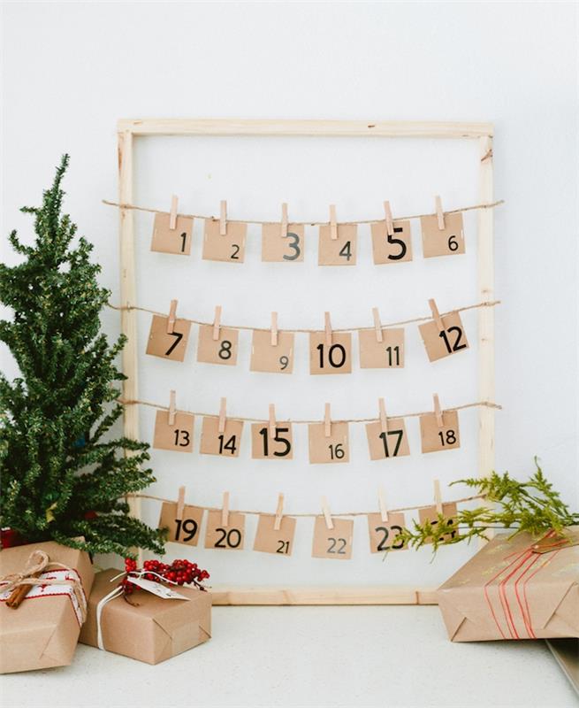 šviesaus medžio rėmas su kraftpopieriaus etiketėmis su juodais skaičiais, kabančiais ant siūlų iš medinių skalbinių segtukų, dekoratyvi mini Kalėdų eglutė ir dovanų paketai