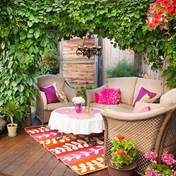 gėlių balkono dekoravimo pavyzdys su ruda sofa ir foteliais, gėlių motyvais dekoruotas kavos staliukas, įvairiaspalvis kilimas, žalios augmenijos arka