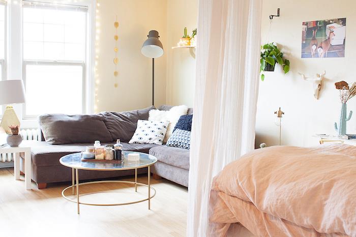 gyvenamojo kambario miegamojo atskyrimas su permatoma šydo stiliaus užuolaida, pilka sofa su dekoratyvinėmis pagalvėmis, lašišos patalynė ant lovos, šviesus parketas, stiklo ir žalvario stalas