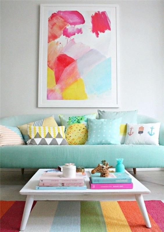 vandens žalios spalvos svetainės išdėstymas, mėlynai žalia sofa, dekoruota spalvingomis pastelinių tonų pagalvėlėmis, įvairiaspalvis dryžuotas kilimas, baltas medinis kavos staliukas, abstrakčios tapybos sienų apdaila