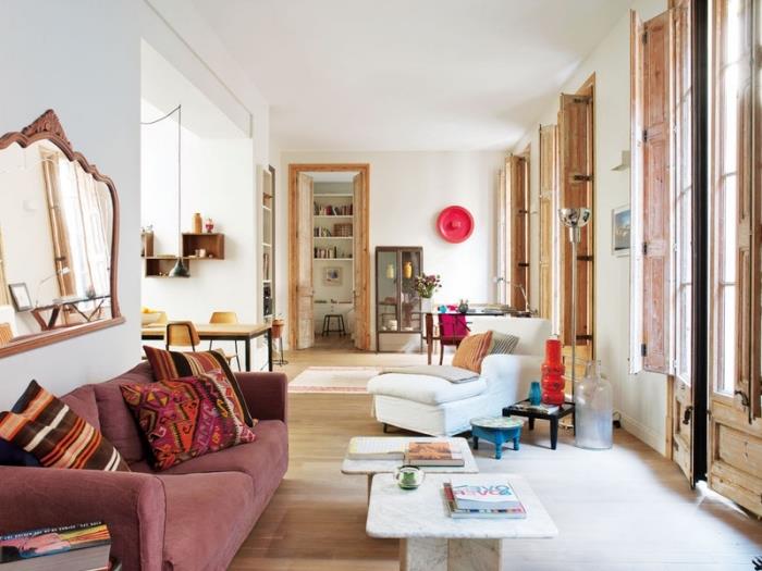 topla notranjost v beli dnevni sobi z lesenimi okni in svetlim parketom, rožnati model kavča, prekrit z etničnimi blazinami