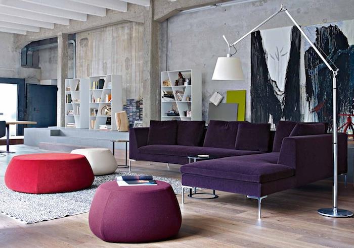 industrijski podstrešni dekor v sodobni dnevni sobi z vijoličnim kavčem, vijoličnimi, belimi in rdečimi mizicami, rdečo preprogo, lahkim parketom, polico za knjige, obrabljeno steno