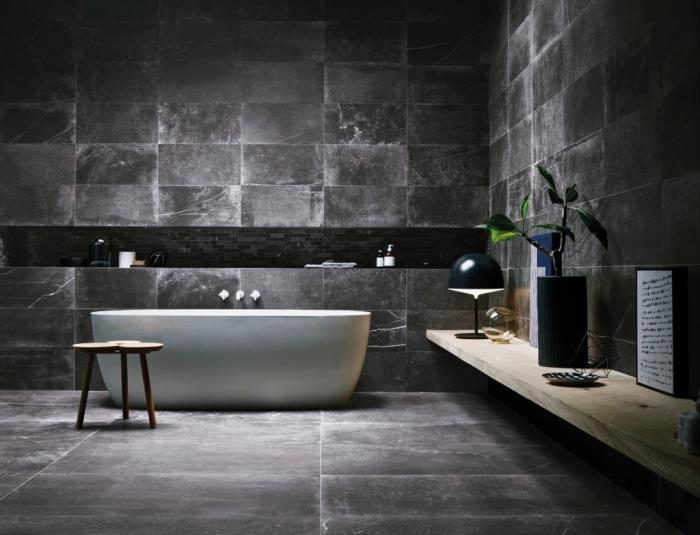 pramoninė vonios kambario apdaila su antracito pilkomis sienomis ir grindimis su balta vonia ir šviesaus medžio lentyna