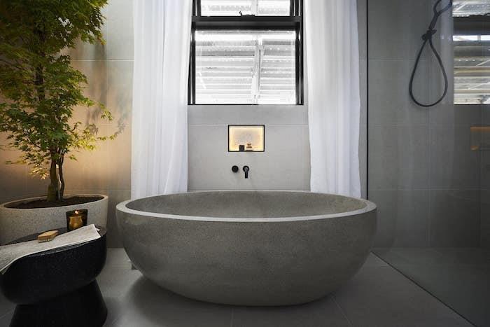 travertino plytelės pilka vonios kambarys natūralaus akmens vonia baltos užuolaidos ir augalas