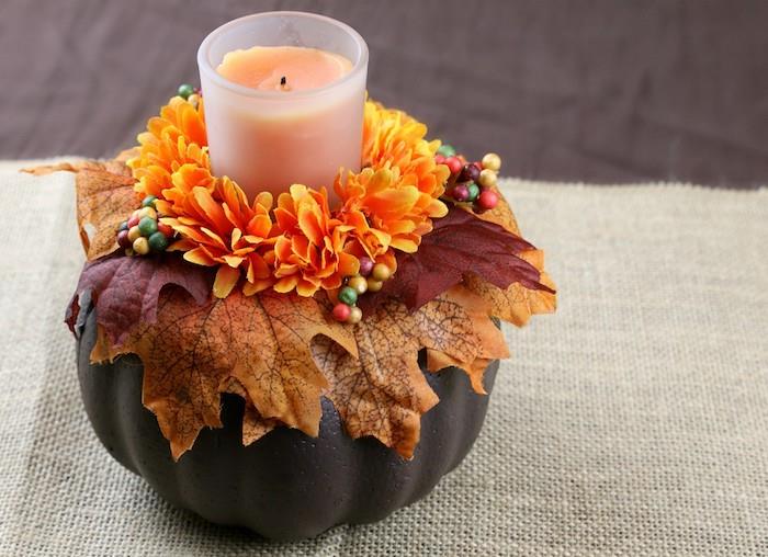 ideja jesenske dekoracije mize z bučo, prebarvano v rjavo barvo z okrasjem jesenskih listov in oranžnimi cvetovi ter svečo na sredini