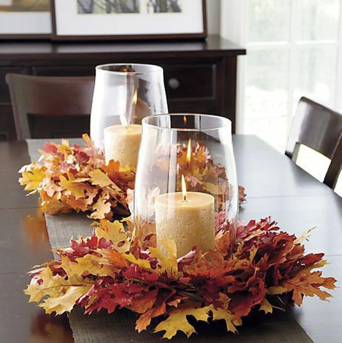 šiltas stalo dekoravimas, uždegtos žvakės, išdėstyti rudeniniai lapai, paprasti stikliniai žvakidės
