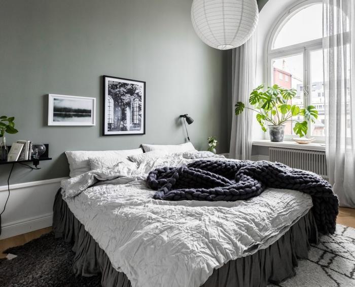 kokoninė lova pilkai baltai, pilkai dažyta siena, balta rutulinė lubų lempa, arkinis langas, maža lentyna