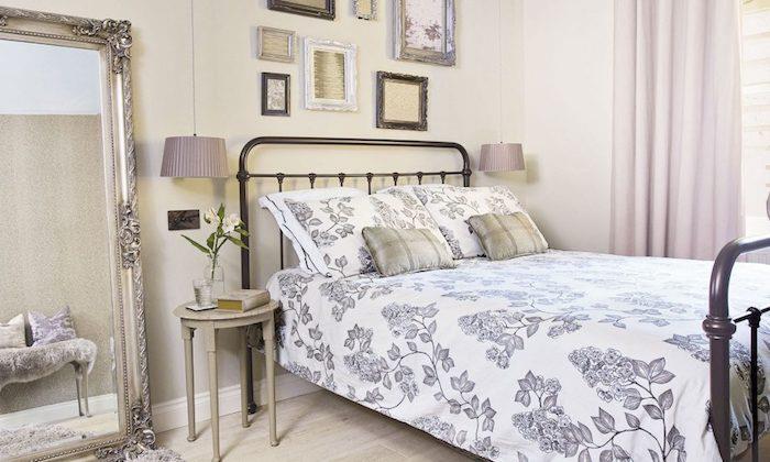 bež spalnica s stensko dekoracijo, baročno ogledalo, kovinska postelja, lahki leseni parket, sivo -belo posteljnina, stenska dekoracija okvirjev