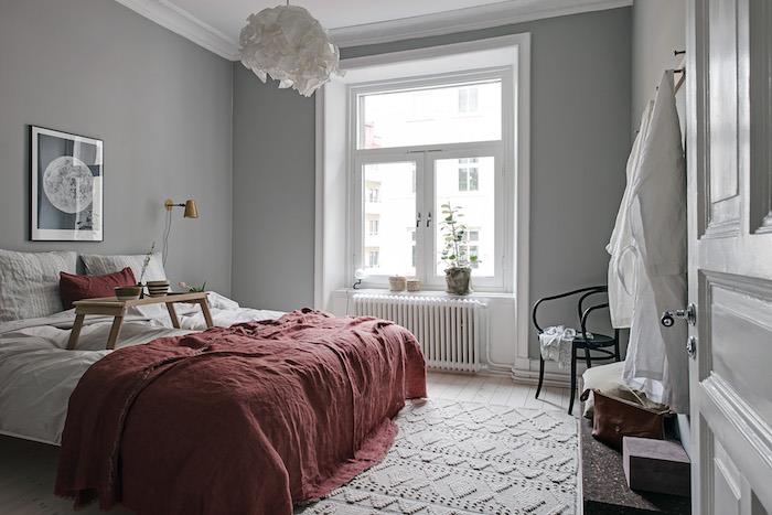 Skandinaviško stiliaus miegamojo apdaila pilka ir balta, raudona lovatiesė, megztas kilimas, balta pakabinama šviesa, šviesaus medžio parketas